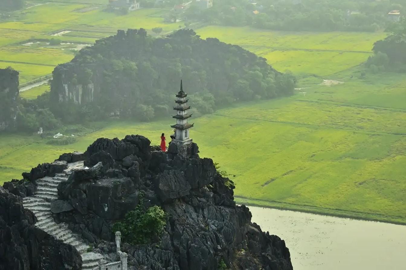 An international traveler standing beside the iconic stone dragon monument at the summit of Hang Mua peak at dawn.  